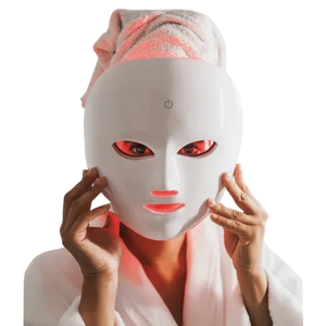 A person wearing a white LED light therapy mask, with red light glowing, and a towel wrapped around their head.