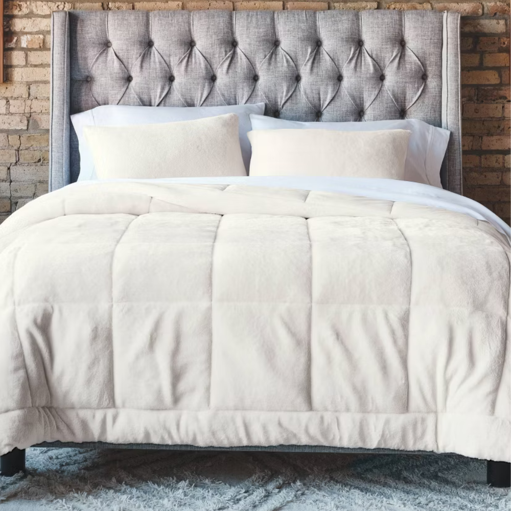 A neatly made bed features a tufted gray headboard, white pillows, and a plush cream comforter, set against a brick wall and atop a fluffy gray rug.