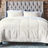 A neatly made bed features a tufted gray headboard, white pillows, and a plush cream comforter, set against a brick wall and atop a fluffy gray rug.