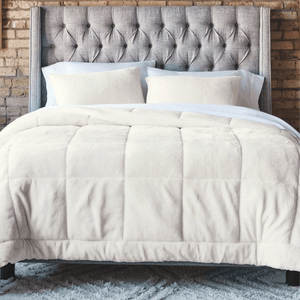 A neatly made bed features a tufted gray headboard, white pillows, and a plush cream comforter, set against a brick wall and atop a fluffy gray rug.