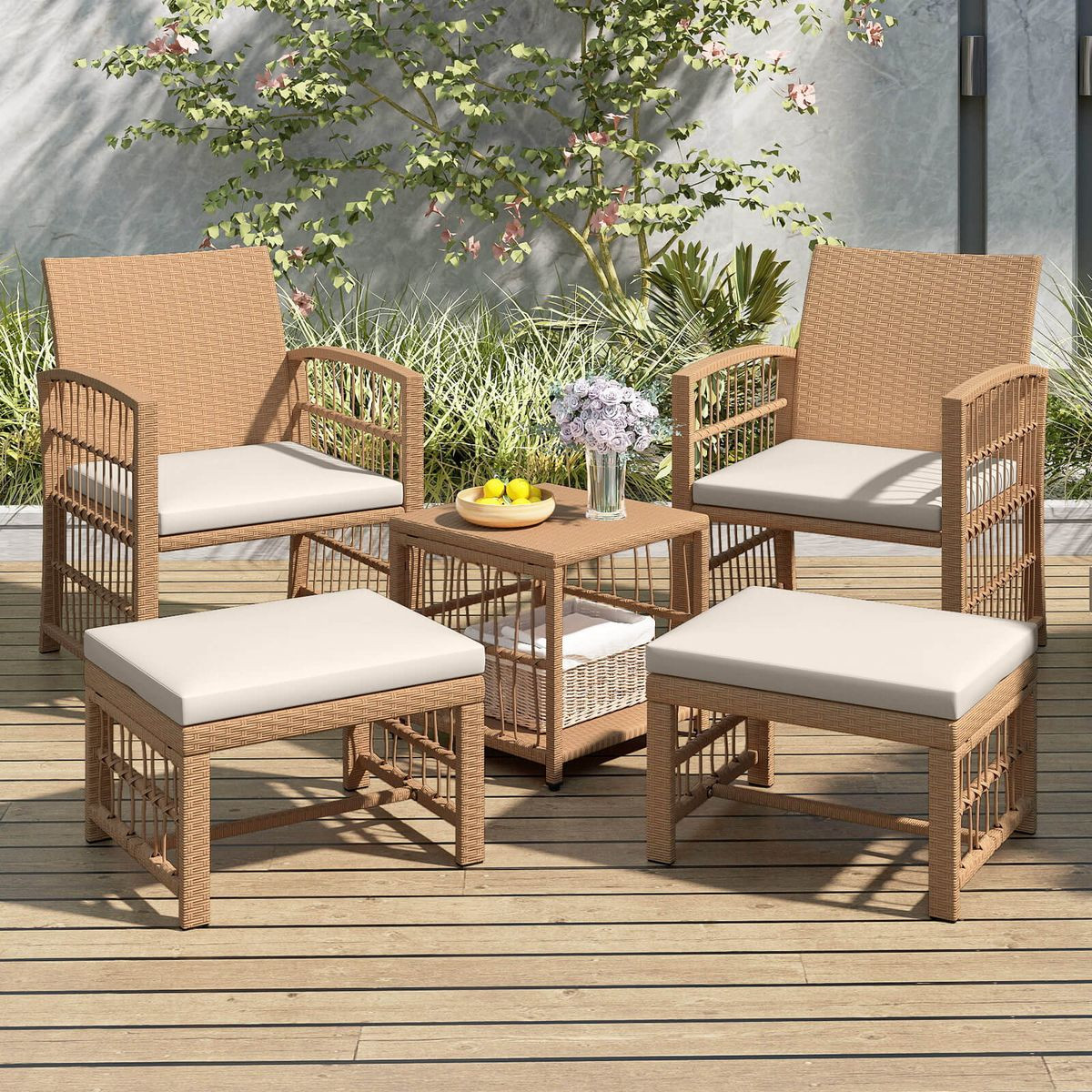 Outdoor patio set with two wicker chairs, two ottomans, and a small table. Cushions are beige, and the table holds a bowl of lemons and a vase with flowers. Surrounded by greenery on a wooden deck.