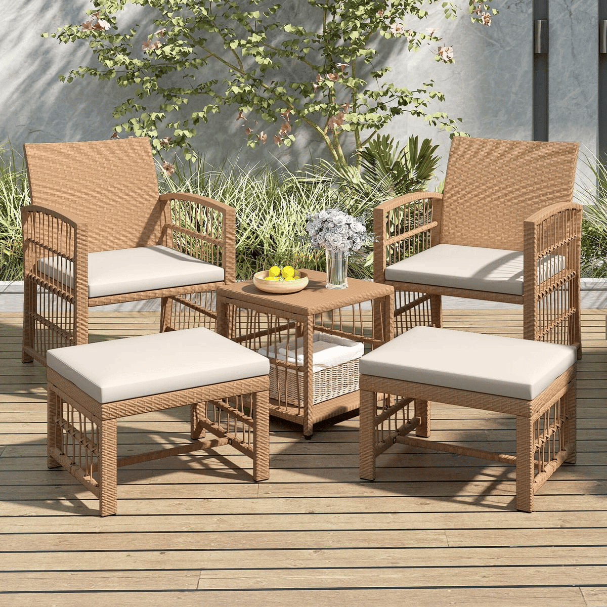 Outdoor patio set with two wicker chairs, two ottomans, and a small table. Cushions are beige, and the table holds a bowl of lemons and a vase with flowers. Surrounded by greenery on a wooden deck.