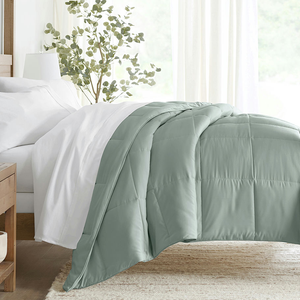 A light green quilted comforter is draped over a bed with white sheets, next to a wooden bedside table and a green plant.