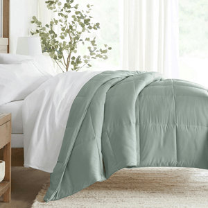 A light green quilted comforter is draped over a bed with white sheets, next to a wooden bedside table and a green plant.