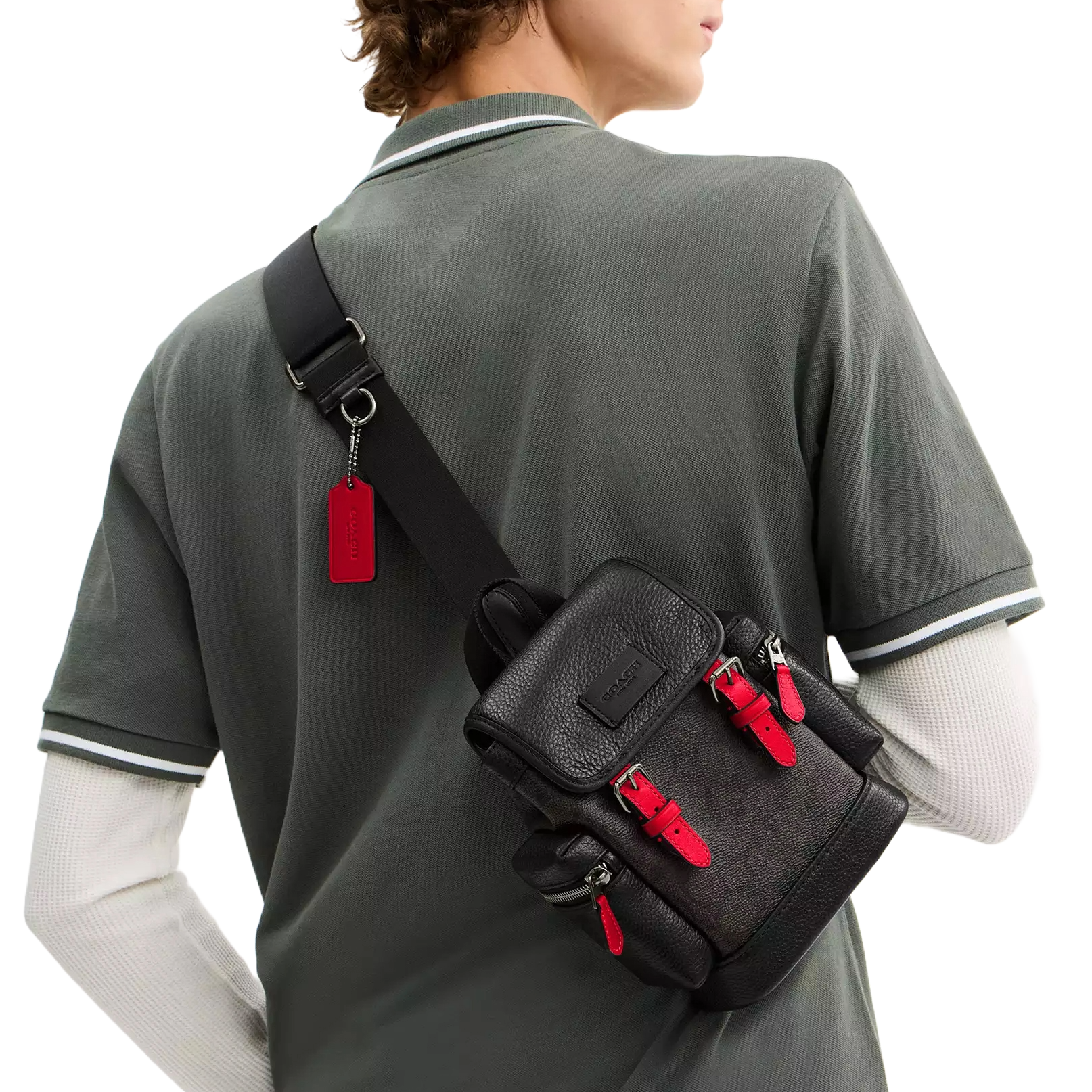 A person is wearing a black leather crossbody bag with red accents and zippers. The bag has multiple compartments and a visible brand tag hanging from the strap.