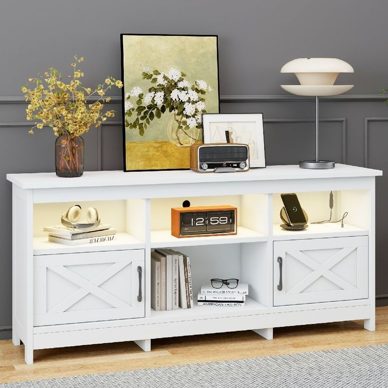 A white sideboard features shelves with LED lighting, displaying books, a clock, a phone, and headphones. The top holds a vase with flowers, artwork, a radio, and a modern lamp.