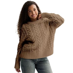 A person wearing a brown cable-knit sweater and jeans, posing with one hand on their chin.