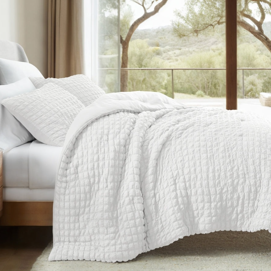 White quilted bedspread and matching pillows on a bed with a light gray upholstered headboard in a room with a large window overlooking greenery.
