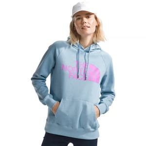 A woman wears a light blue North Face Half Dome Hoodie with a pink logo emblazoned on the front. She pairs it with a white cap and has her hands in the hoodie's front pocket.
