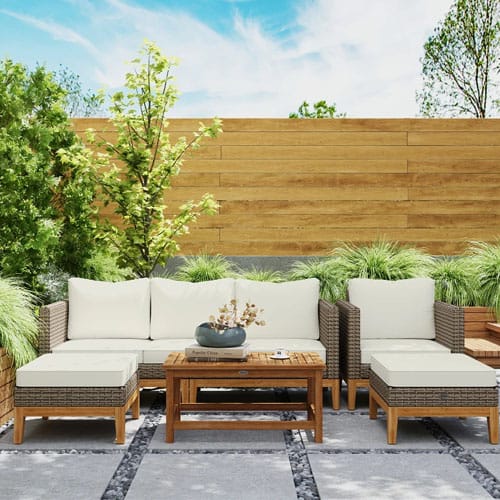 Outdoor rattan furniture set with cream cushions includes a sofa, two ottomans, and a wooden coffee table on a patio with plants and a wooden fence backdrop.