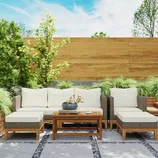 Outdoor rattan furniture set with cream cushions includes a sofa, two ottomans, and a wooden coffee table on a patio with plants and a wooden fence backdrop.