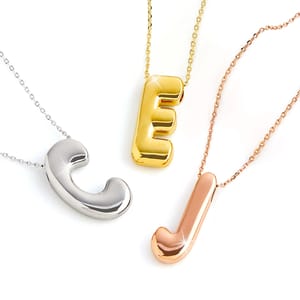 Three bubble letter necklaces are shown with different metallic finishes: a silver 'C', a gold 'E', and a rose gold 'J', each hanging on matching chains.