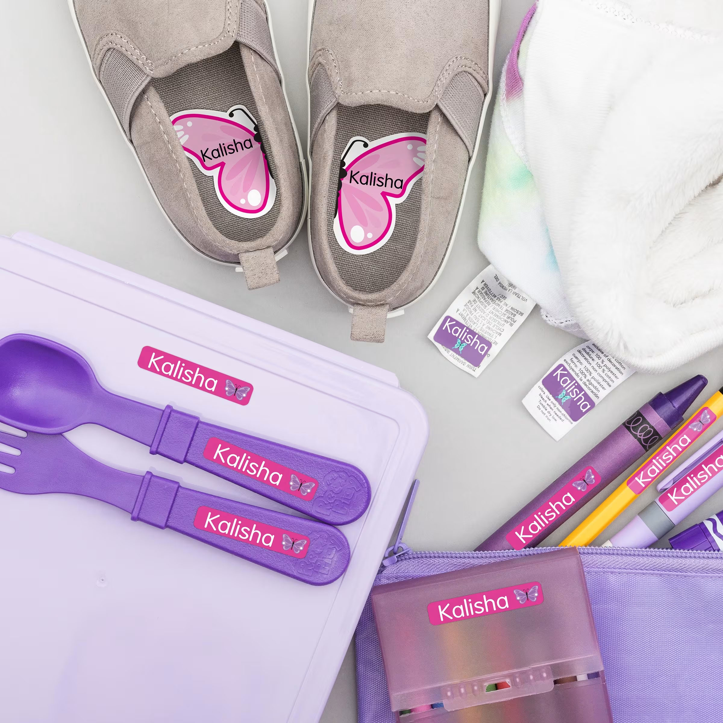 A collection of items labeled \“Kalisha\“ including gray shoes with butterfly insole labels, a purple utensil set, a lavender lunchbox, a fuzzy white garment, and various pencil case supplies, showcasing personalized name labels.