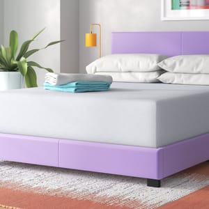 A purple bed frame with a grey mattress is topped with neatly stacked blue and white towels and paired with white pillows. A plant, yellow lamp, and decorative artwork are visible in the background.