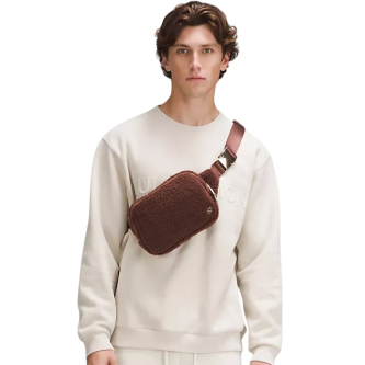 A man wears a cream sweatshirt and carries a brown crossbody bag with a textured finish.