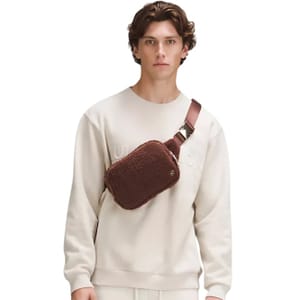 A man wears a cream sweatshirt and carries a brown crossbody bag with a textured finish.