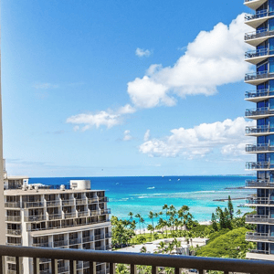 View from a balcony overlooking a coastal cityscape, featuring high-rise buildings, palm trees, and a clear blue ocean under a partially cloudy sky.