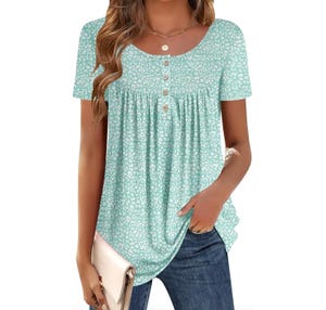 A woman is wearing a light teal short-sleeve top with a white floral pattern and button details on the front, paired with blue jeans and holding a light beige clutch.