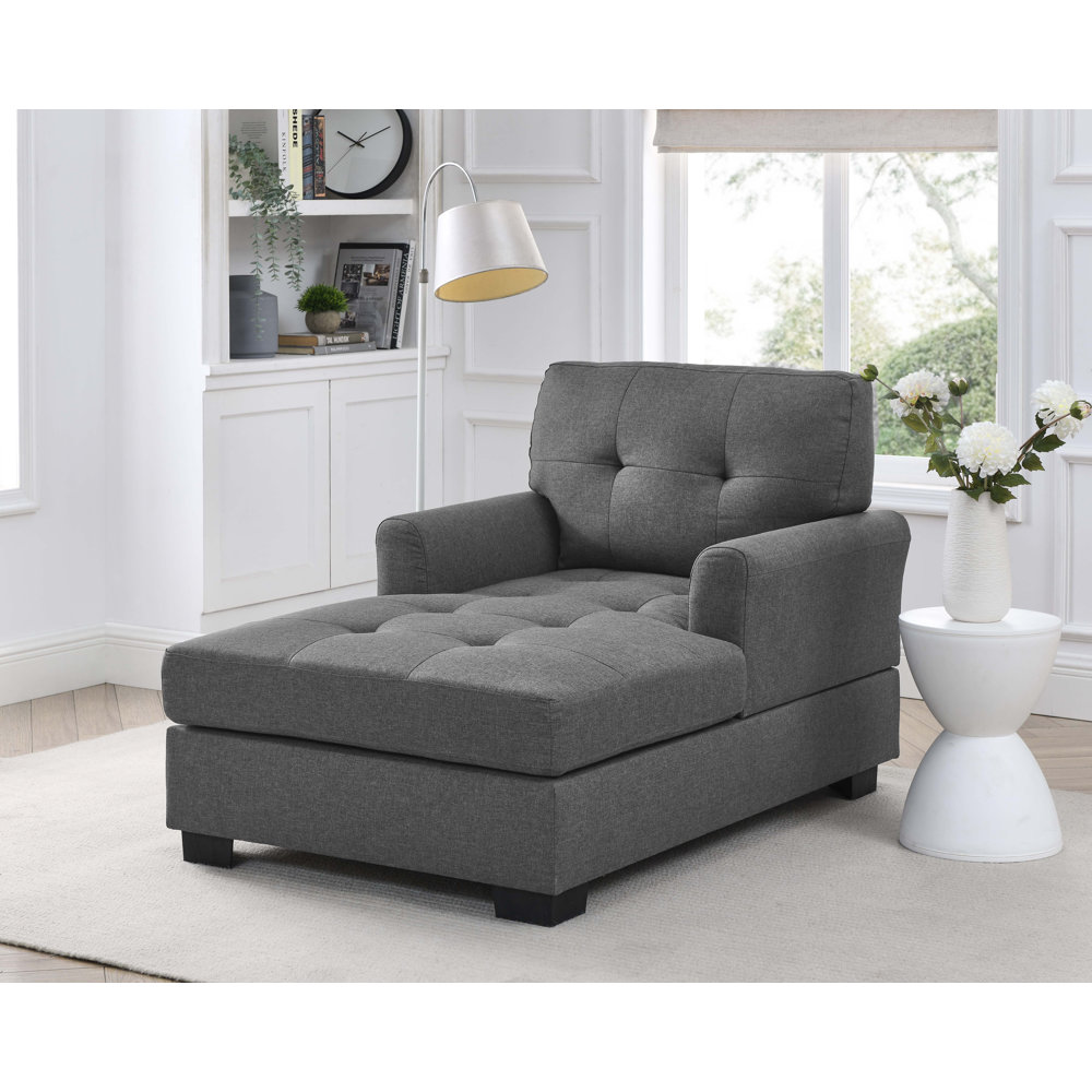 A gray upholstered chaise lounge chair with a cushioned backrest, seat, and an extended ottoman, set in a bright room with a white floor lamp overhead.