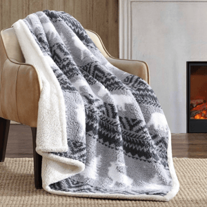 A cozy gray and white fleece blanket with a pattern is draped over a tan leather chair. The setting includes a fireplace and a snowy scene outside the window.