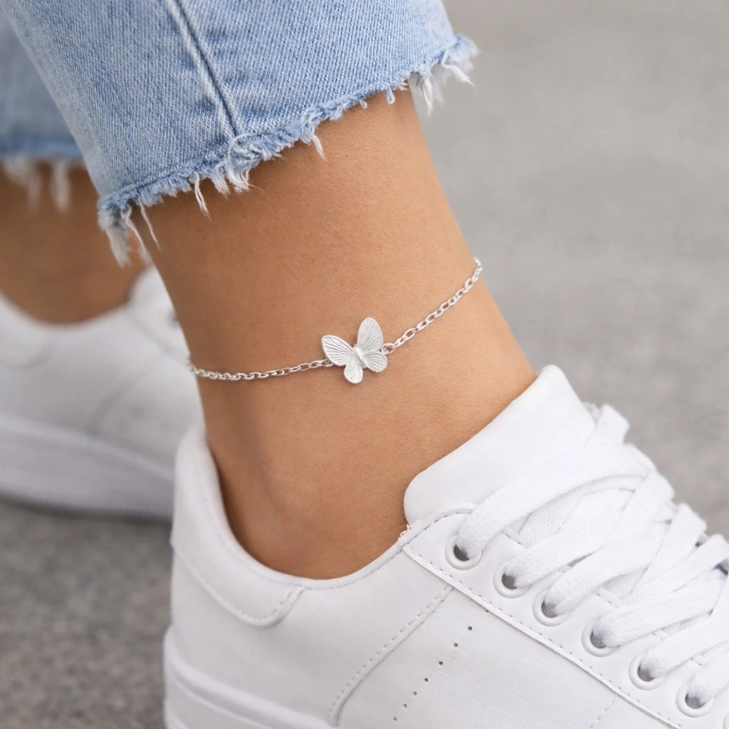 A silver chain anklet features a butterfly charm, worn above a white sneaker with distressed light blue jeans.
