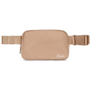 Beige crossbody fanny pack with a large zipper compartment, adjustable strap, and plastic buckle. The name \“Clarisa\“ is printed on the front in white.