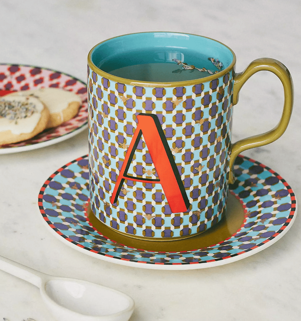 Colorful mug with a geometric pattern and a large red letter \“A\“ on the front, featuring a gold handle and interior. It sits on a matching saucer, accompanied by a decorative cookie plate and a white spoon.