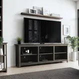 A modern TV stand with a sleek dark wood finish, featuring open shelving and decorative plants. There's a large flat-screen TV above the stand, with additional framed art and books on a wall-mounted shelf above.