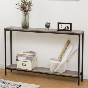 A modern console table with a metal frame and wooden top features a vase with flowers, a date display showing \“Jan 28\“, a decorative blue globe, a box, books, and a basket holding rolled papers. A framed artwork hangs above, and a plant and patterned throw add accents.