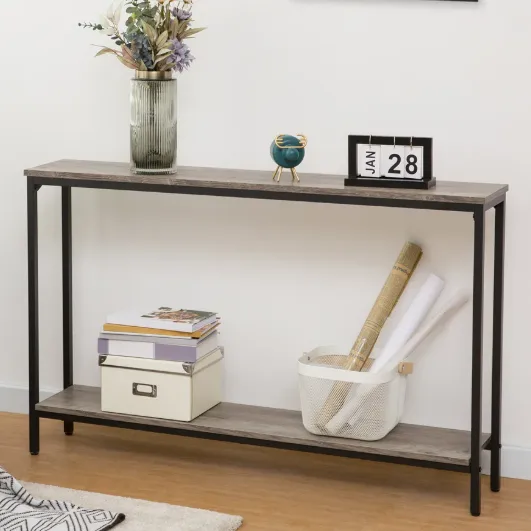 A modern console table with a metal frame and wooden top features a vase with flowers, a date display showing \“Jan 28\“, a decorative blue globe, a box, books, and a basket holding rolled papers. A framed artwork hangs above, and a plant and patterned throw add accents.