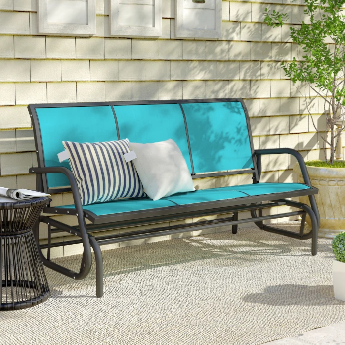 Outdoor patio setup with a turquoise glider bench featuring striped and solid cushions, a wire side table, and potted plants against a shingled wall with decorative framed art.
