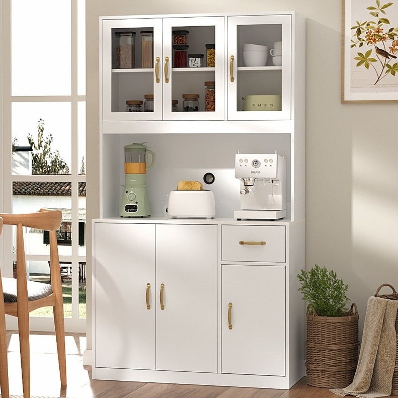 A white kitchen cabinet with gold handles holds a blender, toaster, and espresso machine. Upper glass cabinets display jars and cups. There's a framed botanical print on the wall, chair, and basket with plant and towels nearby.