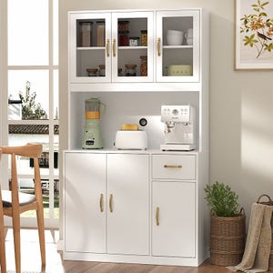 A white kitchen cabinet with gold handles holds a blender, toaster, and espresso machine. Upper glass cabinets display jars and cups. There's a framed botanical print on the wall, chair, and basket with plant and towels nearby.
