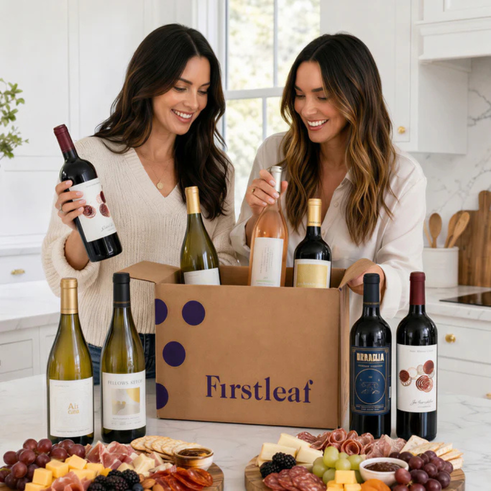 Two women unpack a Firstleaf wine delivery box containing various wine bottles, surrounded by a charcuterie board with cheese, grapes, crackers, and meats.