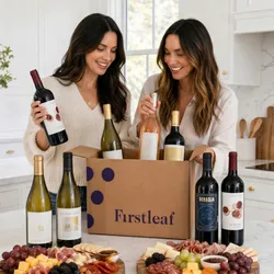 Two women unpack a Firstleaf wine delivery box containing various wine bottles, surrounded by a charcuterie board with cheese, grapes, crackers, and meats.