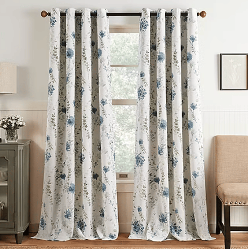 White curtain panels with blue floral patterns hanging on a black rod, set in a cozy room with a cabinet, lamp, and beige chair.