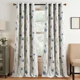 White curtain panels with blue floral patterns hanging on a black rod, set in a cozy room with a cabinet, lamp, and beige chair.