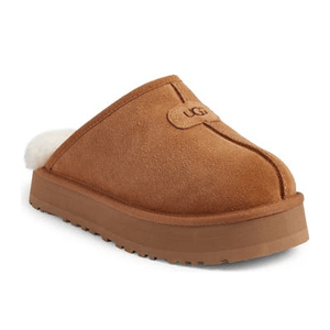 A tan suede slip-on shoe with a thick sole and white fleece lining, featuring the UGG logo on the upper.