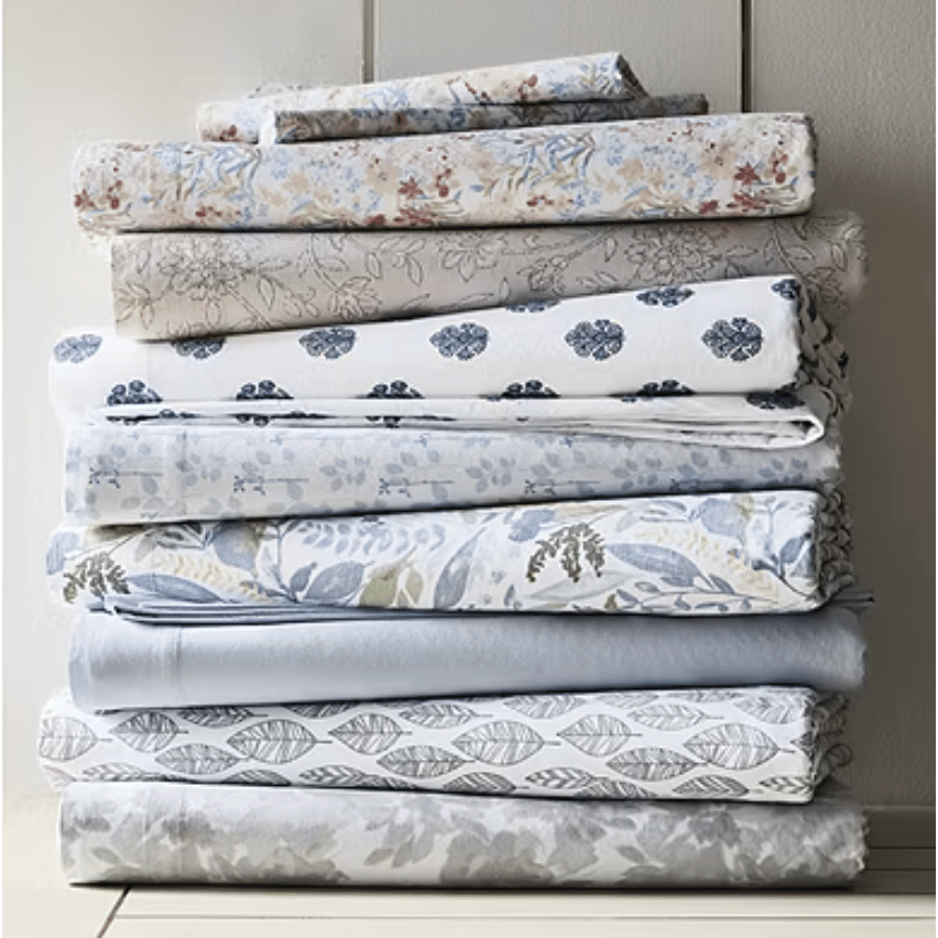 A stack of folded bedsheets featuring various floral and leaf patterns in soft pastel colors, including blue, gray, and beige tones.