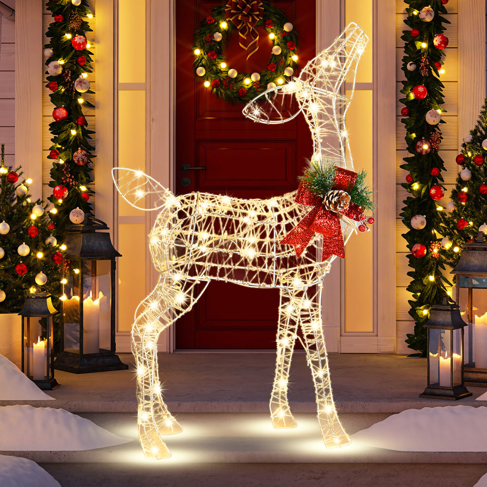 A festive lighted reindeer decoration is positioned between two snow-dusted, lantern-flanked columns adorned with garlands and a wreath on the door.