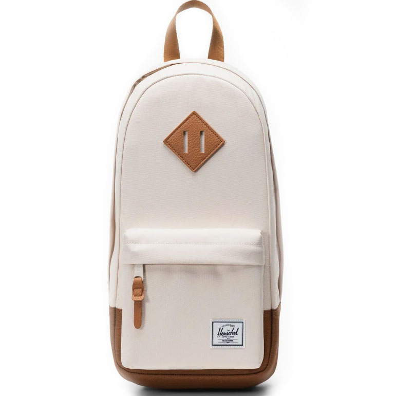 A cream-colored Herschel backpack featuring a brown faux-leather diamond-shaped accent, a front pocket with a key clip, and the brand's white label on the lower front.
