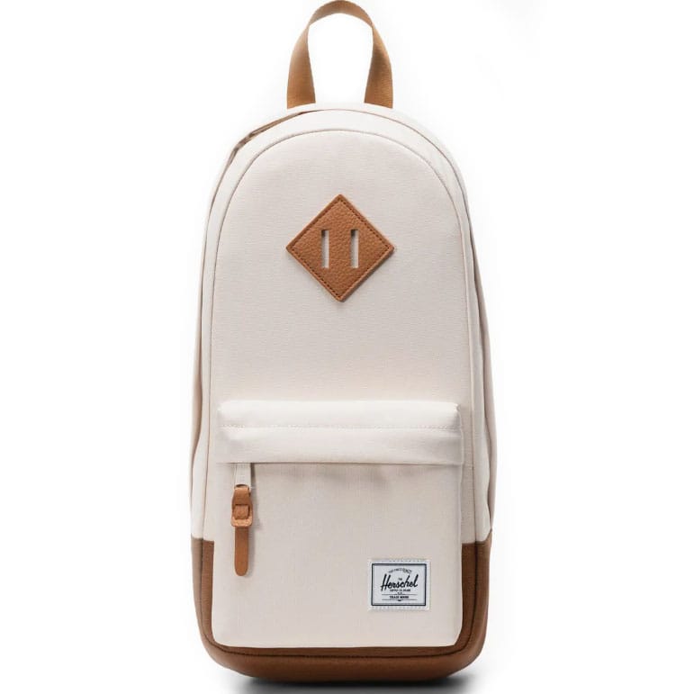 A cream-colored Herschel backpack featuring a brown faux-leather diamond-shaped accent, a front pocket with a key clip, and the brand's white label on the lower front.