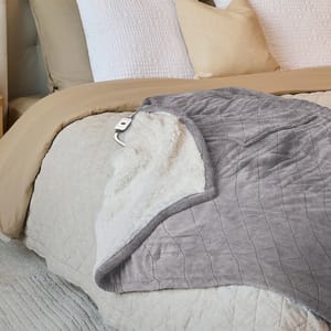 A heated reversible Sherpa electric throw blanket featuring one side of plush Sherpa and the other of smooth fabric, equipped with an attached controller for temperature adjustment, draped over a bed layered with beige and white bedding.