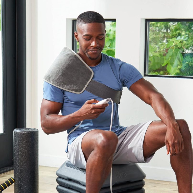 The man is using a wearable heating pad on his shoulder while seated on exercise steps, holding a controller. A foam roller is nearby.