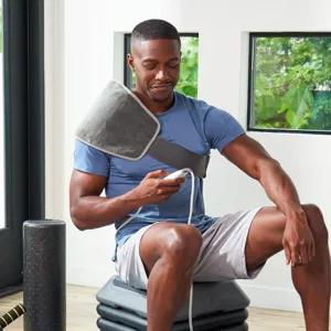 The man is using a wearable heating pad on his shoulder while seated on exercise steps, holding a controller. A foam roller is nearby.