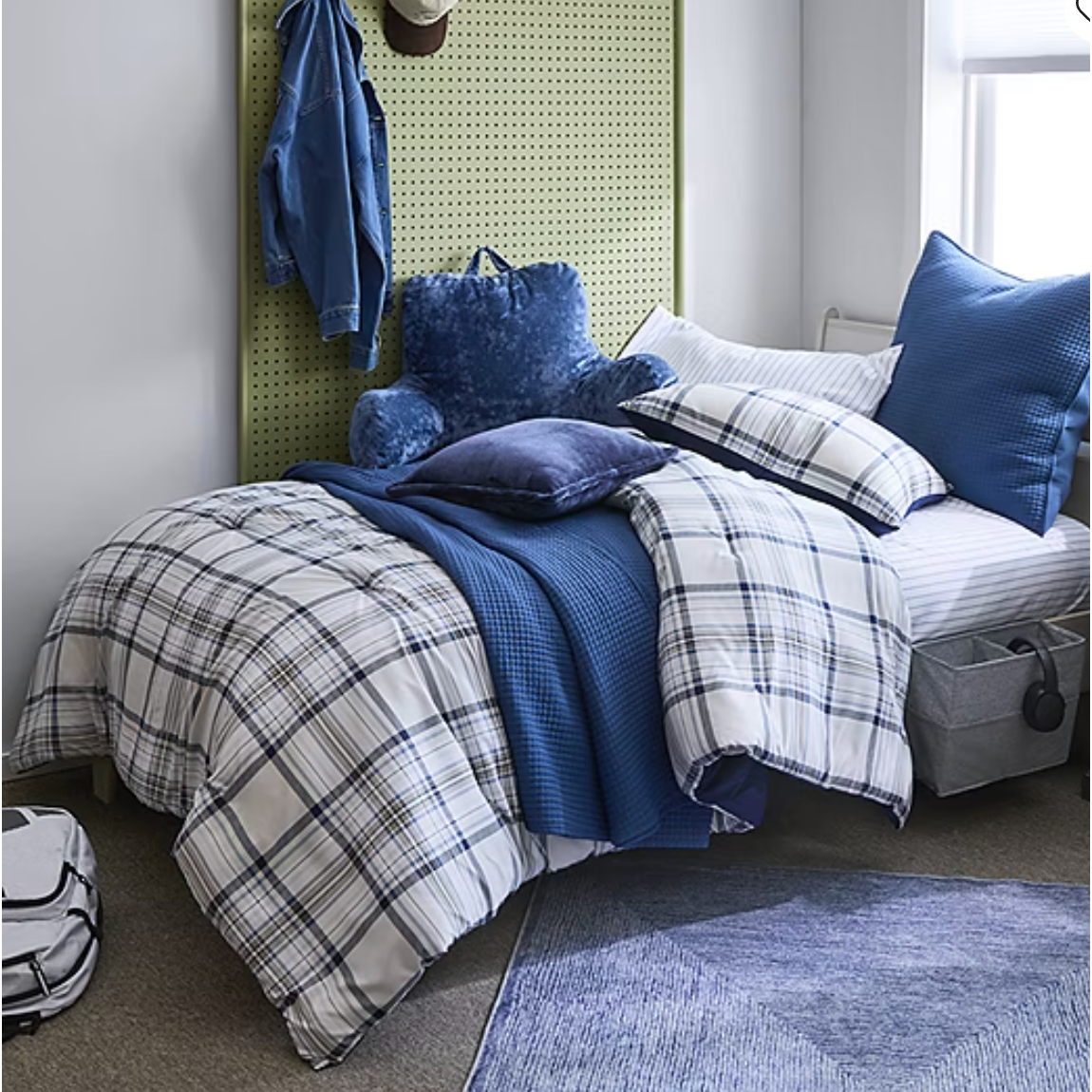 A cozy bedroom setup featuring a bed with blue and plaid bedding, a plush blue backrest pillow, a green pegboard headboard with a denim jacket and hat, and a floor rug. A backpack and storage bins are placed nearby.