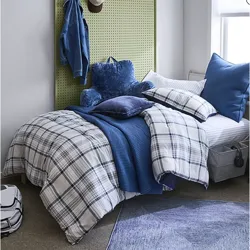 A cozy bedroom setup featuring a bed with blue and plaid bedding, a plush blue backrest pillow, a green pegboard headboard with a denim jacket and hat, and a floor rug. A backpack and storage bins are placed nearby.