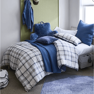 A cozy bedroom setup featuring a bed with blue and plaid bedding, a plush blue backrest pillow, a green pegboard headboard with a denim jacket and hat, and a floor rug. A backpack and storage bins are placed nearby.