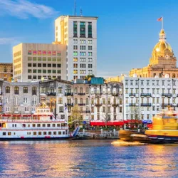 A riverboat and a moving tugboat are in the foreground of a scenic riverfront cityscape, featuring historic multi-story buildings and a dome-topped structure under a clear blue sky.
