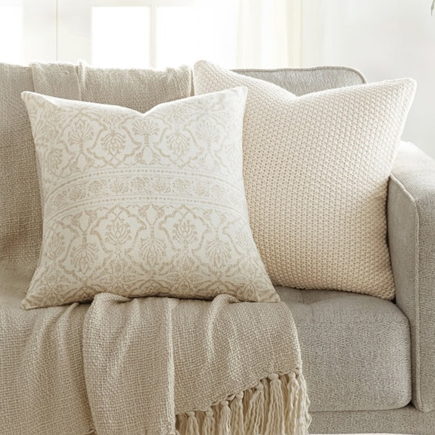 A beige sofa is adorned with two decorative pillows and a fringed throw blanket in neutral tones. A small side table holds a potted plant, adding a touch of greenery to the setting.
